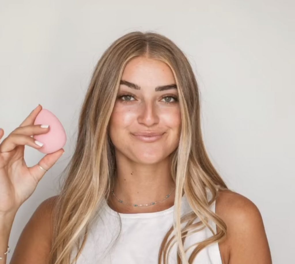 How to use a makeup sponge in 3 simple steps by popular blogger and seint artist, Kelly Snider. Image of a blonde model holding a pink seint perfector sponge. 