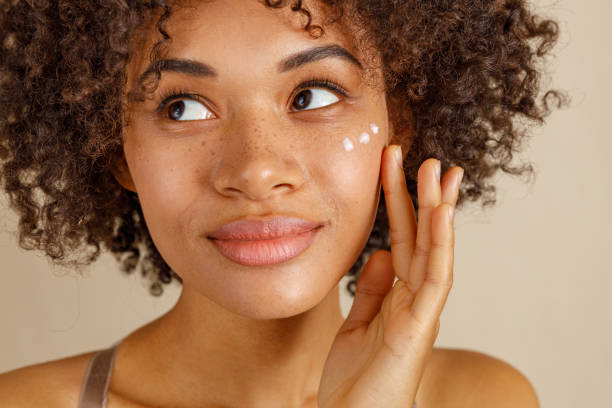 Charming young lady looking at side with dots of face cream on her cheek, isolated on beige color background